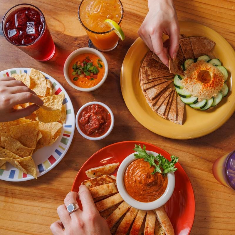 Overhead spread of hummus, pita, chips, dips and drinks, with hands reaching to share.